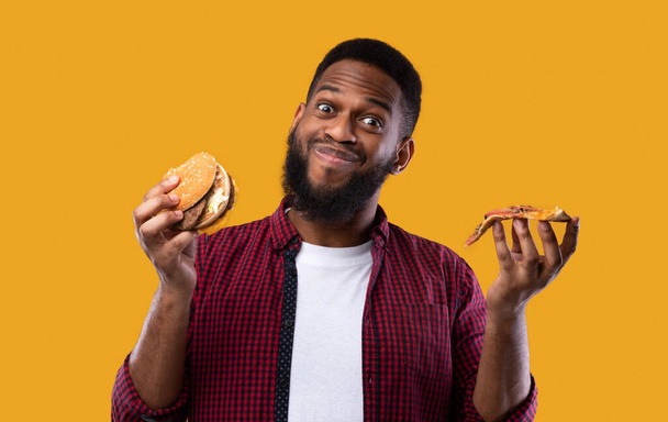 Chico negro feliz sosteniendo hamburguesa y pizza sobre fondo amarillo - Foto, Imagen