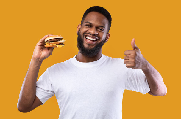 africano chico holding hamburguesa gestos pulgares-up en amarillo estudio fondo - Foto, imagen