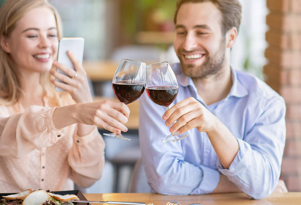 Νεαρή γυναίκα κάνει φωτογραφία του ρομαντικού τοστ με clinking ποτήρια κρασιού κατά τη διάρκεια εορταστική δείπνο στο καφέ, επιλεκτική εστίαση - Φωτογραφία, εικόνα