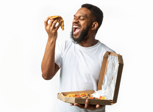 Głodny czarny facet jedzący kawałek pizzy stojący nad białym tle - Zdjęcie, obraz