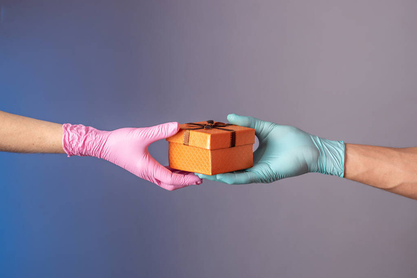A man's hand in a blue glove is giving a gift box to a woman's hand in pink glove on a blue and gray background. Concept of a lifestyle in a pandemic. - Fotó, kép