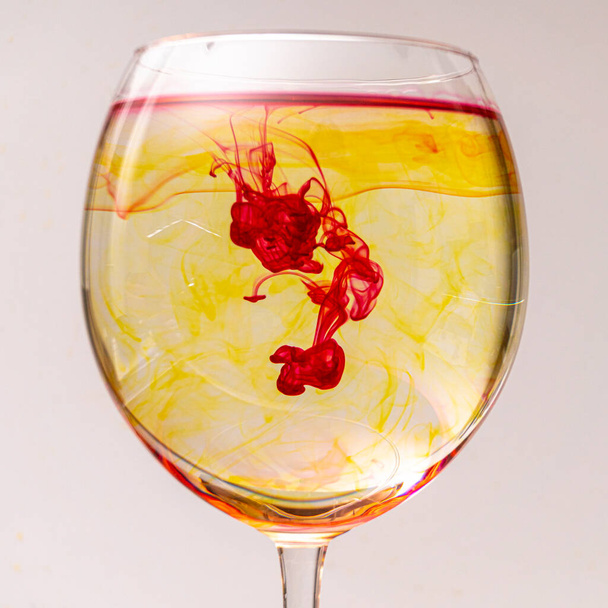 Tropfen farbiger Tinte faellt in ein Glas mit Wasser und ergibt dort ein buntes schlieren artiges Muster. - Foto, Imagem