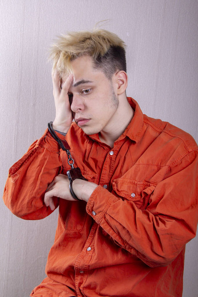 Ein Teenager in Handschellen hält seinen Kopf mit den Händen vor einem grauen Hintergrund. Kopfschmerzen bei jugendlichen Straftätern in Quarantäne, Strafbarkeit. Mitglieder krimineller Banden während der Coronavirus-Zeit - Foto, Bild