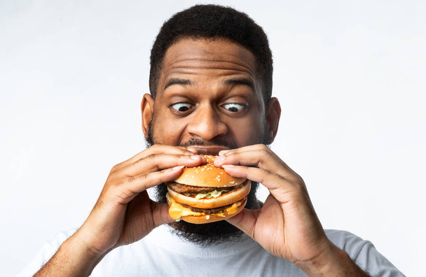 Lustiger hungriger afrikanischer Kerl isst Burger auf weißem Hintergrund - Foto, Bild