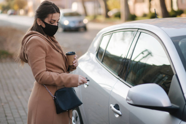 Kobieta w czarnej masce trzyma przy samochodzie mankiet z kawą i szuka kluczyków. Otwarty samochód. Maska protekcjonistyczna w kwarantannie. Cov-19 - Zdjęcie, obraz