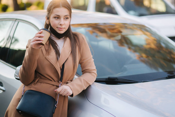Beautiful young woman in black face mask stand by the car and drink coffee. Happy young woman have a break. Female pulled off mask for drinking cappucino outdoors - Foto, Bild