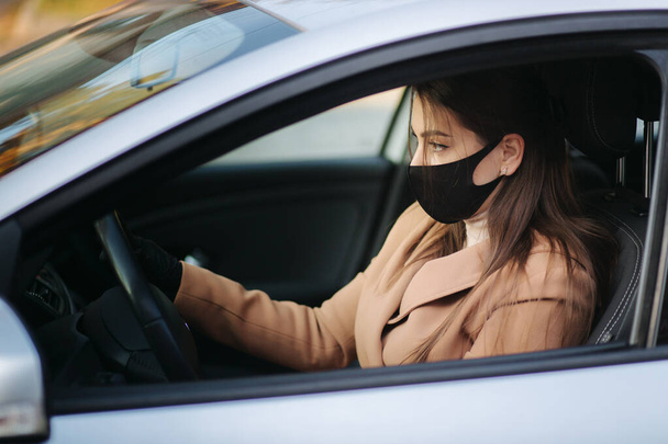 Красивая молодая девушка в маске сидит в машине и надевает защитные перчатки. Защитная маска от ковида-19, водитель на городской улице во время вспышки коронавируса - Фото, изображение