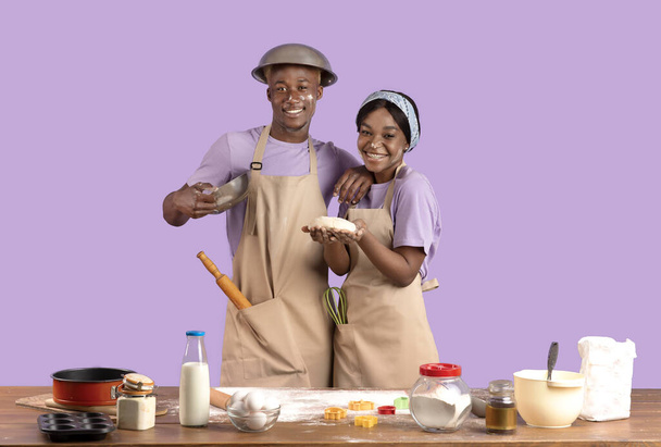 Retrato de una joven pareja negra con caras sucias horneando juntas, sosteniendo masa para galletas festivas sobre fondo violeta - Foto, imagen