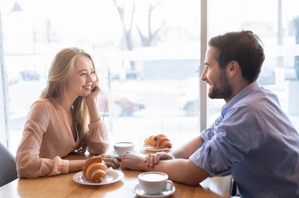 Positiivinen nuori pari aamiaista yhdessä kahvilassa, istuu lähellä ikkunaa ja nauttia keskustelusta - Valokuva, kuva