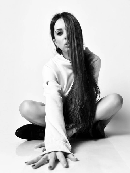 Young beautiful woman with long hair in oversized hoodie and boots sitting on floor, reaching for camera in sexy pose - Фото, изображение