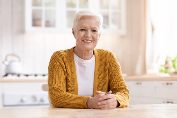 Retrato de una alegre anciana sentada en la cocina - Foto, Imagen