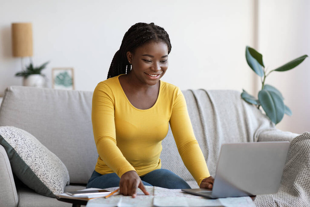 Улыбающаяся черная леди с помощью ноутбука и карты дома, планируя будущие путешествия - Фото, изображение