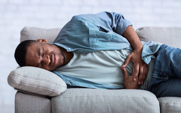 African Guy Suffering From Abdominal Pain Lying On Sofa Indoor - Valokuva, kuva