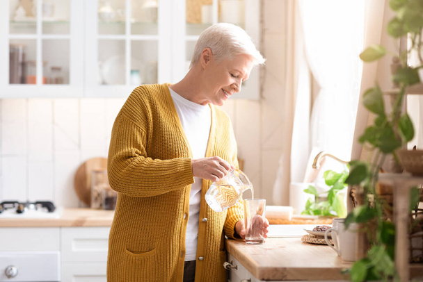 Atractiva abuela vertiendo agua en la cocina en casa - Foto, Imagen
