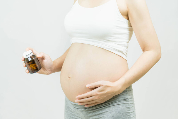 Selective focus of pregnant woman with Medicine bottle in Hand Touching Belly Standing Over Gray  Background,Healthy millennial women taking antioxidant medicine vitamins, healthcare concept. - Photo, Image