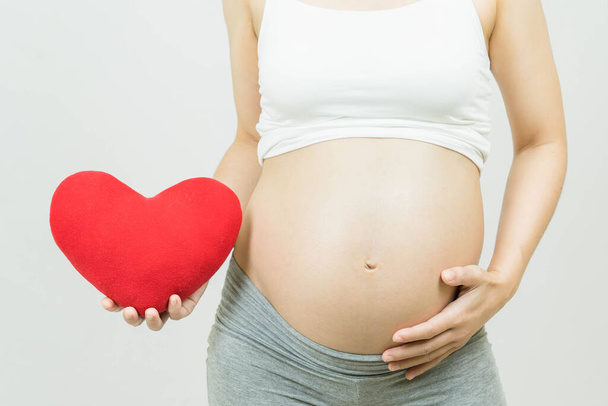 Selective focus of a tummy of a pregnant woman with red heart on hand isolated on gray background, body part, happy pregnancy time, new life concept - Foto, Imagem