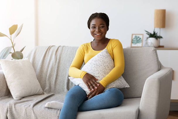 Día en casa. Mujer negra sonriente sentada en un sofá cómodo con almohada - Foto, imagen