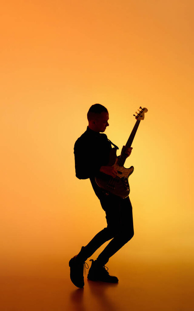 Silhouette of young caucasian male guitarist isolated on orange gradient studio background in neon light - Foto, Imagen