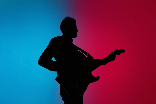 Silhouette of young caucasian male guitarist isolated on blue-pink gradient studio background in neon light - Φωτογραφία, εικόνα