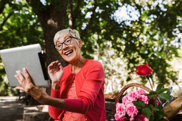 Herrlich lächelnde kaukasische Seniorin mit blonden kurzen Haaren sitzt auf Bank im Park und macht Videotelefonie mit digitalem Tablet. - Foto, Bild