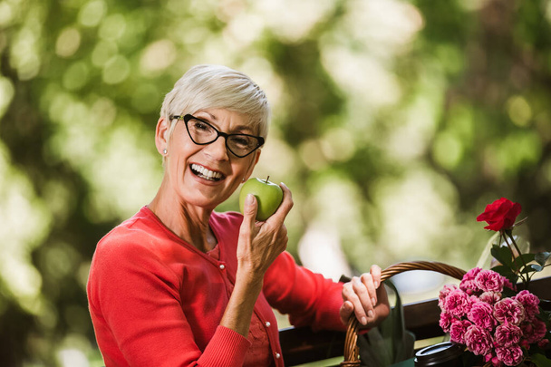 Mujer mayor de aspecto saludable con pelo gris sosteniendo manzana afuera en el parque - Foto, imagen