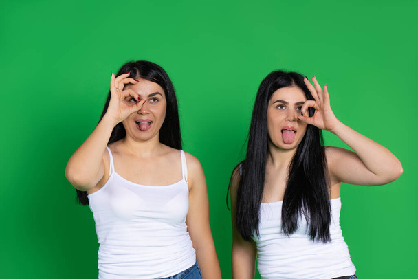 Young twin girls gesturing okay make faces with tongues out on a green background with space. High quality photo - Foto, afbeelding