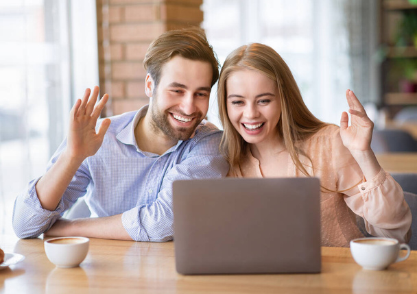 Молодая пара, общающаяся онлайн на ноутбуке в кафе, машущая в камеру - Фото, изображение