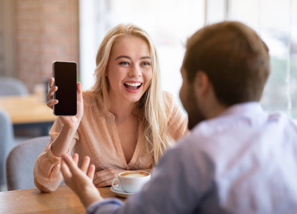 Joven alegre mostrando su teléfono inteligente novio con pantalla vacía en la cafetería, maqueta para el diseño - Foto, imagen