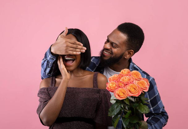 Amante hombre negro sorprendiendo a su novia con ramo de rosas, cubriendo sus ojos sobre fondo de estudio rosa - Foto, imagen