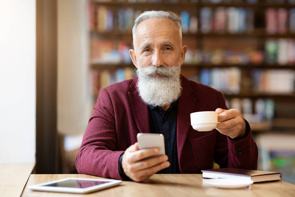 Neşeli gri saçlı yaşlı adam kahve içiyor ve telefon kullanıyor. - Fotoğraf, Görsel