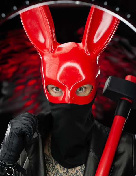 un hombre con una máscara de conejo rojo en una capa de cuero con un hacha - Foto, Imagen