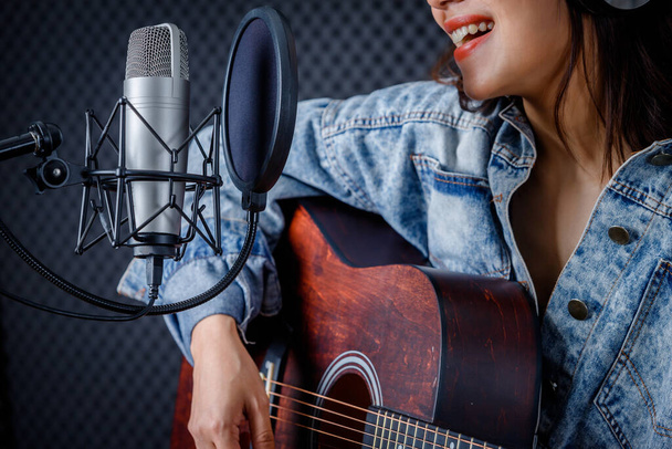 Happy cheerful pretty smiling of portrait of young Asian woman vocalist Wearing Headphones with a Guitar recording a song front of microphone in a professional studio - Valokuva, kuva