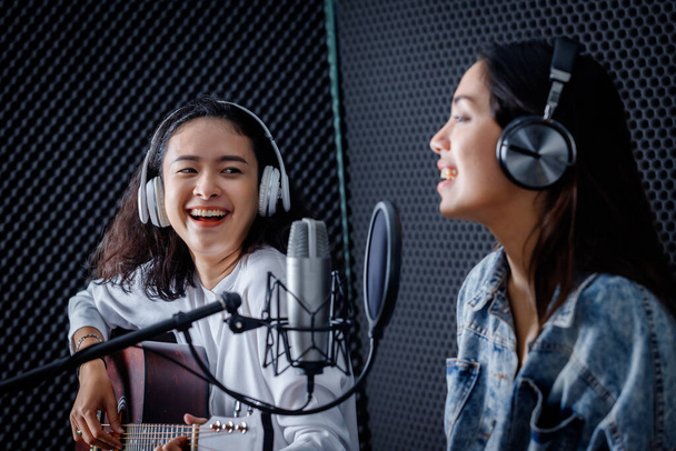 Happy cheerful pretty smiling of portrait Two young Asian woman vocalist Wearing Headphones with a guitar recording a song front of microphone in a professional studio - Photo, image