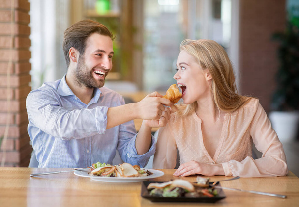 Milující mladý muž krmení chutné croissant na jeho přítelkyně v útulné kavárně - Fotografie, Obrázek