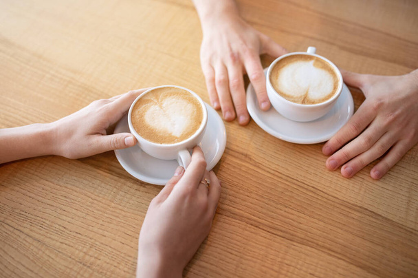 カフェでテーブルの上にハート型の泡のコーヒーを持っている若いカップルの作物のビュー - 写真・画像