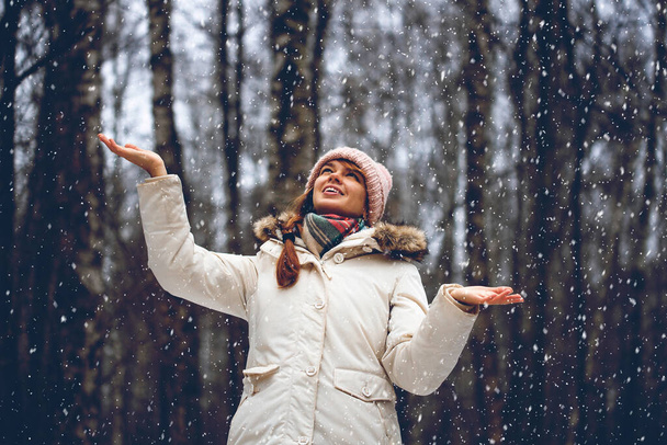 Young woman in a winter park in snowfall. Woman having fun on winter nature. Winter portrait of funny woman in woolen hat and warm scarf throwing snow in winter park, flying snowflakes. - Photo, Image