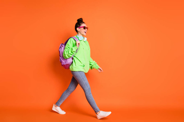 Full length photo of walking young lady dressed green sweatshirt backpack looking empty space isolated bright orange color background - Фото, зображення