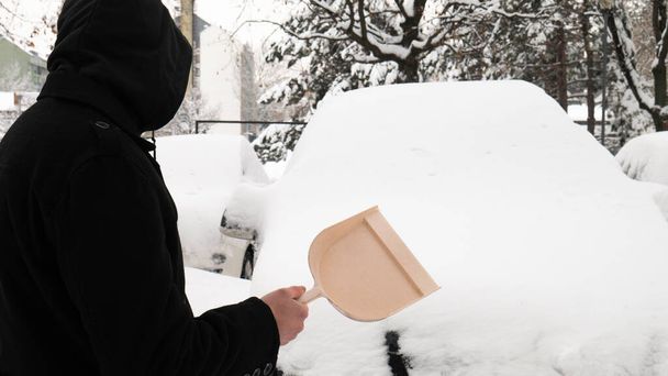 Een man met een kap en een plastic schop klaar om de sneeuw uit de auto te halen in de ochtend voor het werk. - Foto, afbeelding