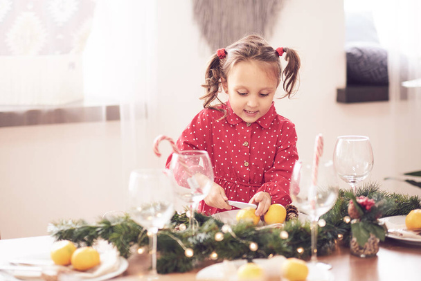 Cute curly toddler girl standing at a Christmas dinner table settling the dishes preparing to celebrate Xmas Eve a decorated dining room with tree and lights - Фото, зображення