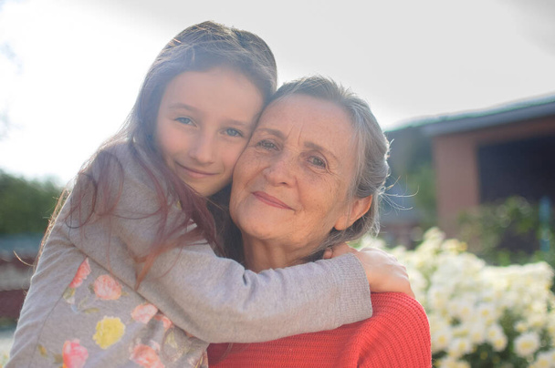 Starsza babcia z siwymi włosami w czerwonym swetrze z wnuczką przytulają się w ogrodzie, a w słoneczny dzień na świeżym powietrzu, dzień matki - Zdjęcie, obraz
