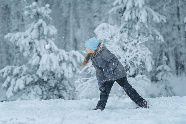 dziewczyna zimą w szarej kurtce, niebieski kapelusz z długimi blond włosami w czarnych spodniach na tle pokrytego śniegiem lasu z drzewami, sosnami, jodłami, gra śnieżki, puszysty biały śnieg, dryfuje - Zdjęcie, obraz