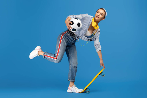 Cheerful young woman with yellow penny or skateboard, soccer ball and headphones posing over blue background. - Foto, imagen