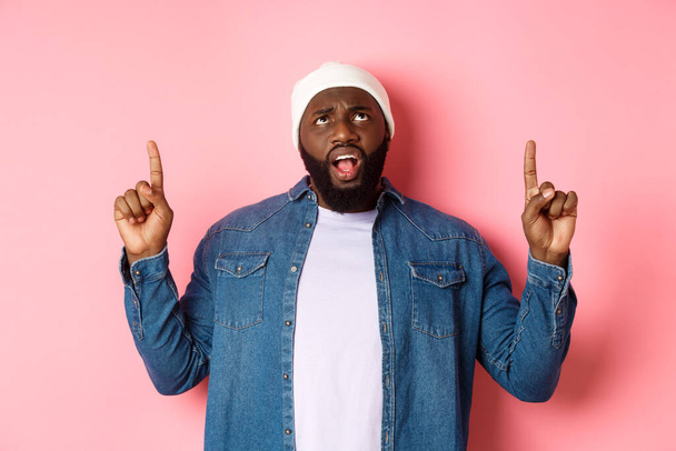 Displeased african american guy in beanie, pointing fingers up and staring at camera bothered, complaining, standing over pink background - Valokuva, kuva