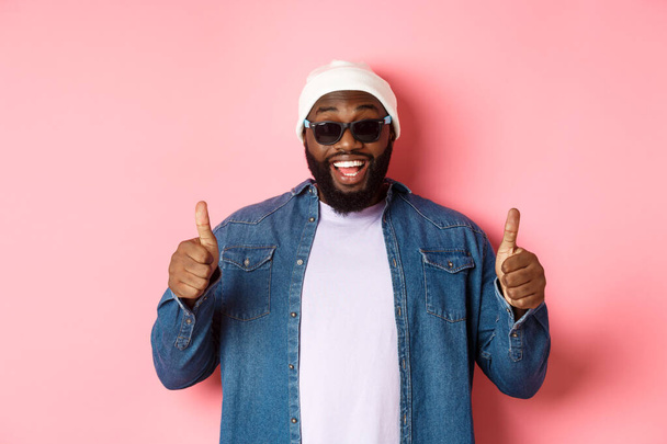Tevreden Afrikaans-Amerikaanse hipster man in beanie en zonnebril tonen duimen-up, goedkeuren en prijzen goede keuze, glimlachen blij, roze achtergrond - Foto, afbeelding