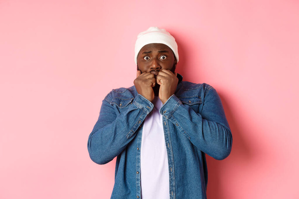 Scared Black guy trembling from fear, biting fingernails and staring at camera, standing over pink background - Photo, Image