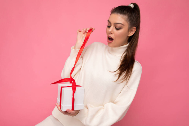 Photo shot of pretty positive surprised young brunet woman isolated over colourful background wall wearing trendy outfit look holding gift box and looking at present box with red ribbon - Photo, Image