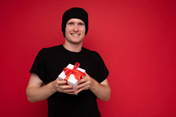 Fénykép lövés jóképű pozitív mosolygós fiatalember elszigetelt felett piros háttér fal visel fekete kalap és fekete póló gazdaság fehér ajándék doboz piros szalaggal, és nézi a kamera - Fotó, kép