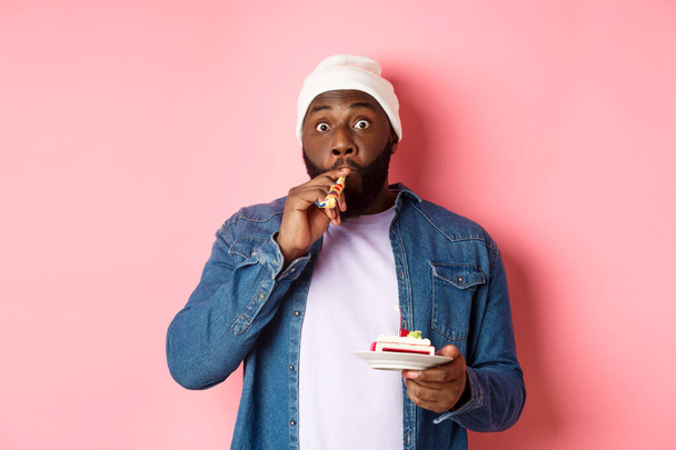 Happy Black hipster celebrating birthday, blowing party whistle, holding bday cake with candle, standing over pink background - Photo, image