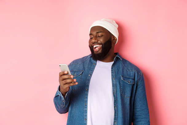 Technológia és online vásárlási koncepció. Boldog fekete szakállas férfi olvasás üzenet és mosolygós, okostelefon rózsaszín háttér - Fotó, kép