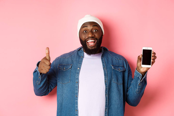 Online-Shopping und Technologie-Konzept. Glücklicher afrikanisch-amerikanischer Hipster-Mann zeigt Daumen hoch und mobilen Bildschirm, lobt App, steht über rosa Hintergrund - Foto, Bild